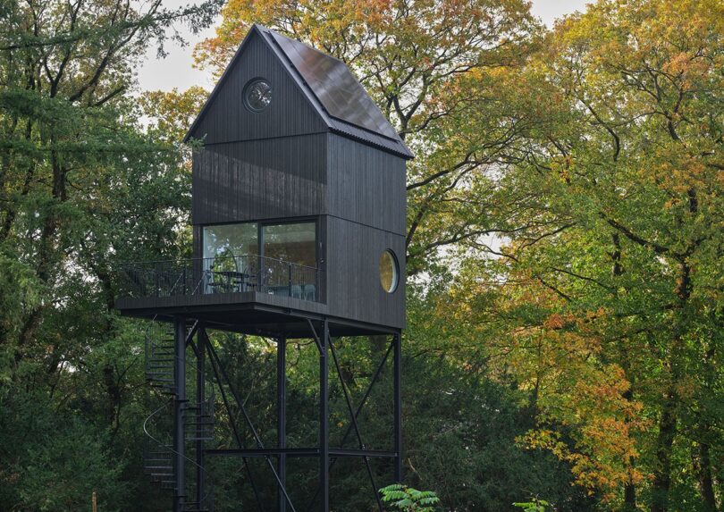 A tall, modern black treehouse on stilts with large windows and a spiral staircase, surrounded by trees with autumn foliage