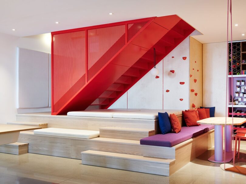 Modern interior with a bold red staircase, wood paneling, a cushioned seating area with colorful pillows, and a rock climbing wall feature