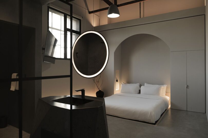 Minimalist bedroom with a modern design features a large bed, a circular mirror with backlighting, a sleek sink, and subdued lighting against neutral grey walls