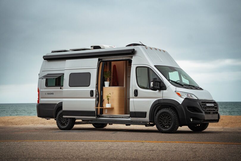 A silver camper van parked by the beach with its side door open, revealing a minimalist wooden interior and a potted plant