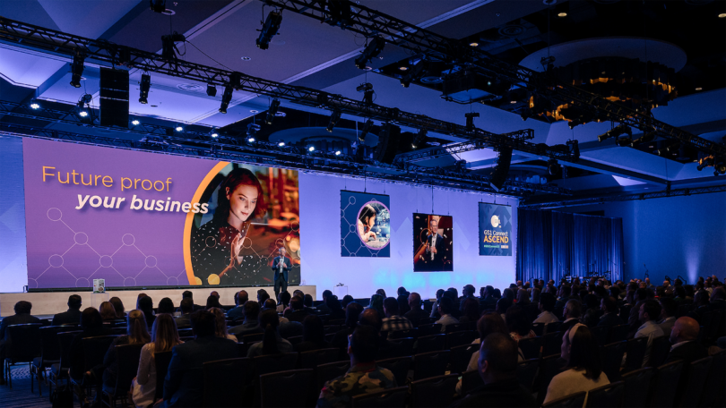 A presenter speaks to an audience at a conference. The stage displays "Future proof your business" with related images and graphics