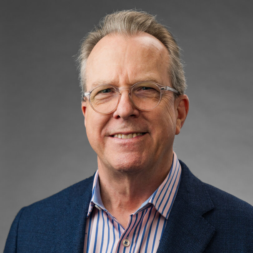 A person wearing glasses, a blue jacket, and a striped shirt, smiling against a plain gray background