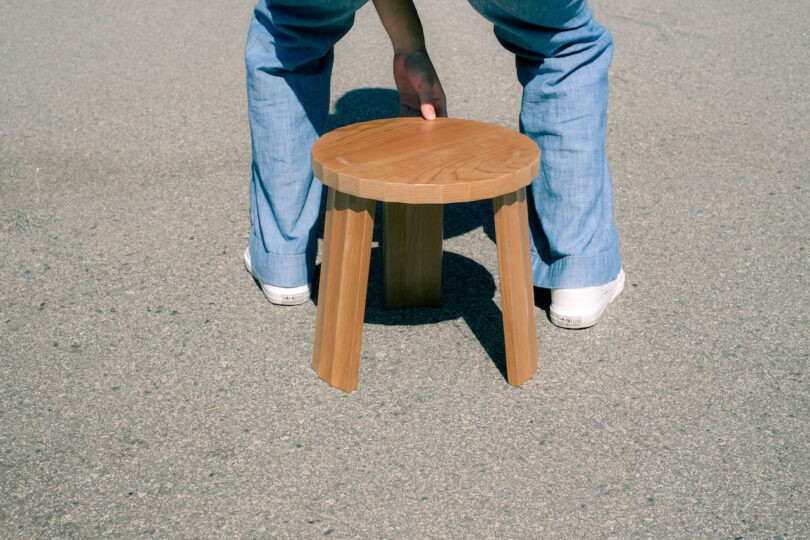 A person in jeans and white shoes kneels to place a wooden stool on a paved ground