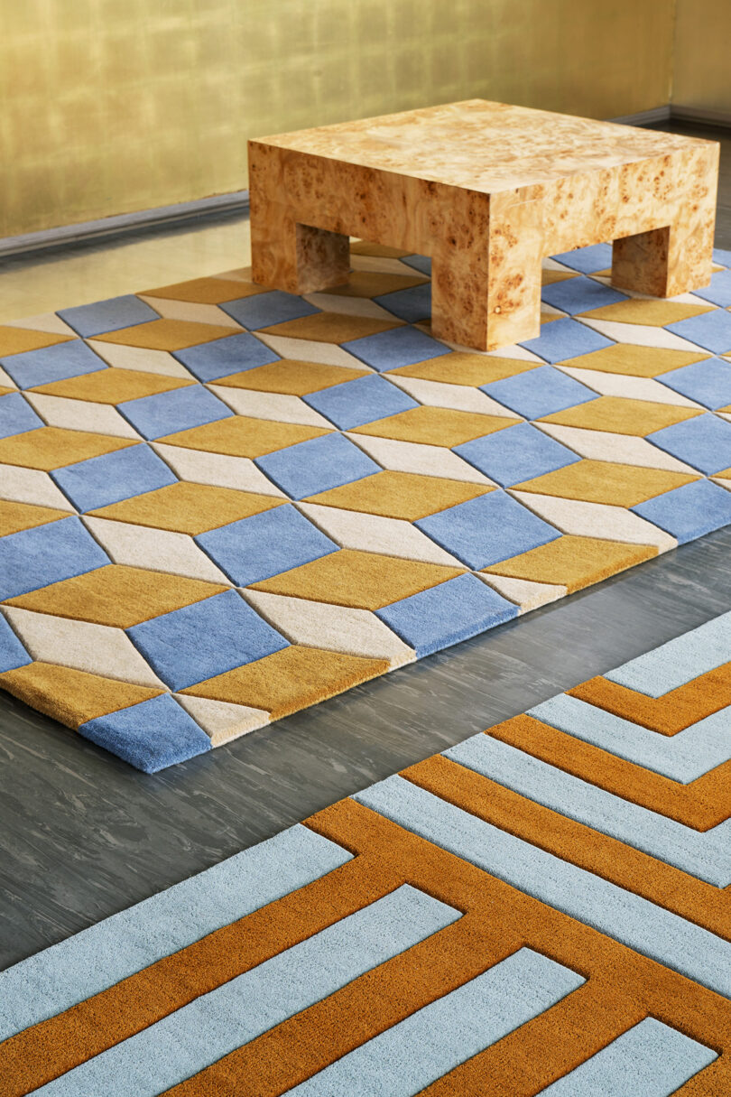 A geometric rug with blue, beige, and yellow shapes lies beneath a square wooden table. Nearby, an artwork from the 10 Years Unfolded LAYERED collection features another rug with blue and orange patterns partially visible in the foreground.