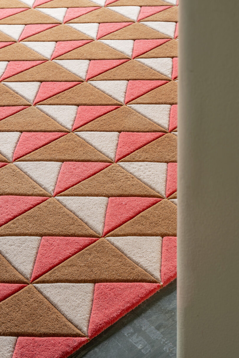 A geometric-patterned rug showcases brown, pink, and beige triangles on a floor, partially obscured by a wall, embodying the timeless elegance of LAYERED design.