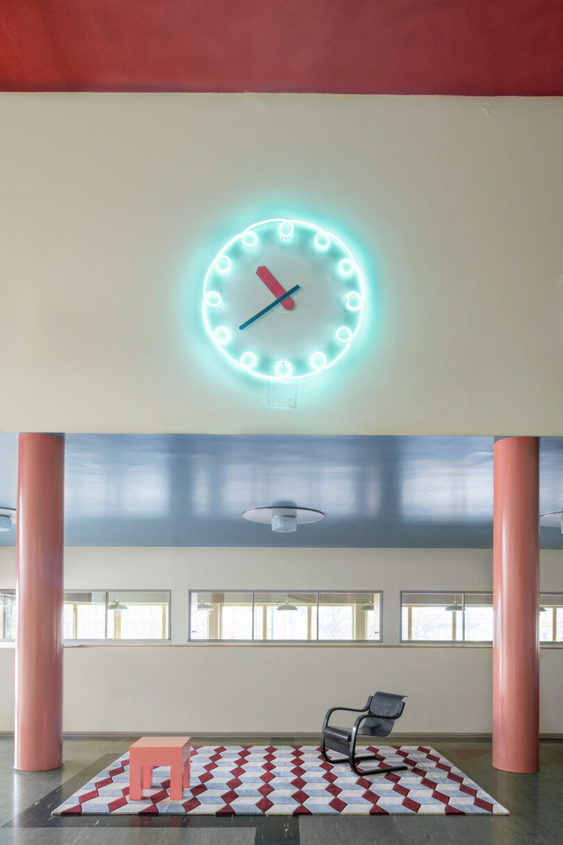 A neon-lit clock casts a glow above a room where layers of 10 years unfold, featuring an orange table, black chair, and geometric carpet.