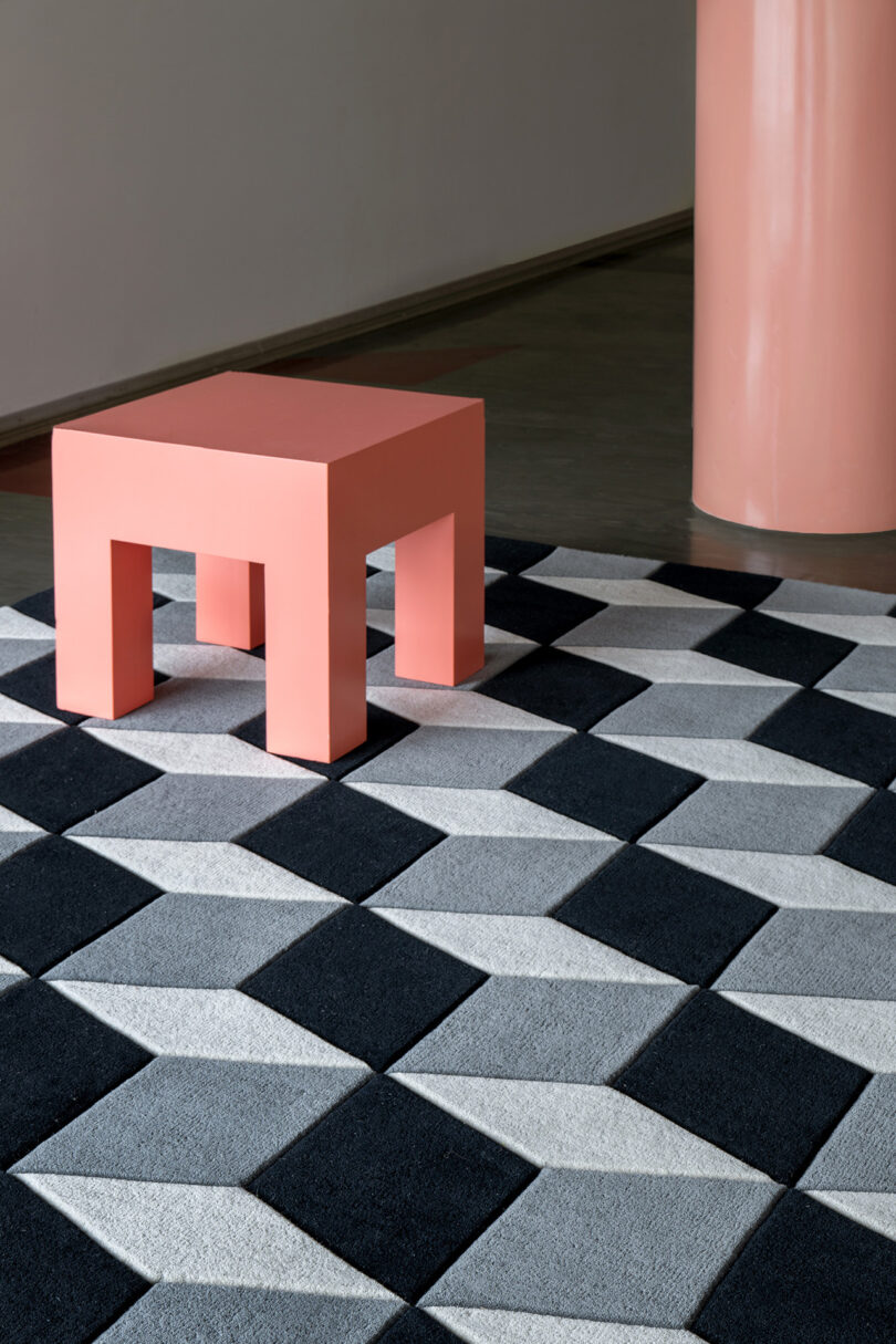 A pink square stool graces a geometric black, white, and gray patterned rug, echoing the artful elegance LAYERED has curated over 10 years, next to a pink cylindrical column.