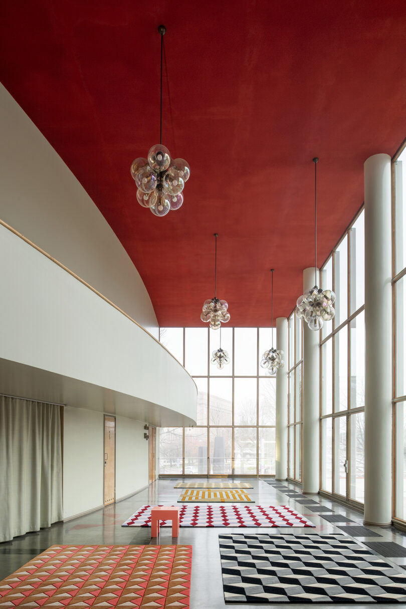 The room boasts a striking red ceiling, large windows, and patterned rugs, creating a LAYERED visual appeal. Cluster pendant lights illuminate the space, where white pillars stand beside elegant curtains.