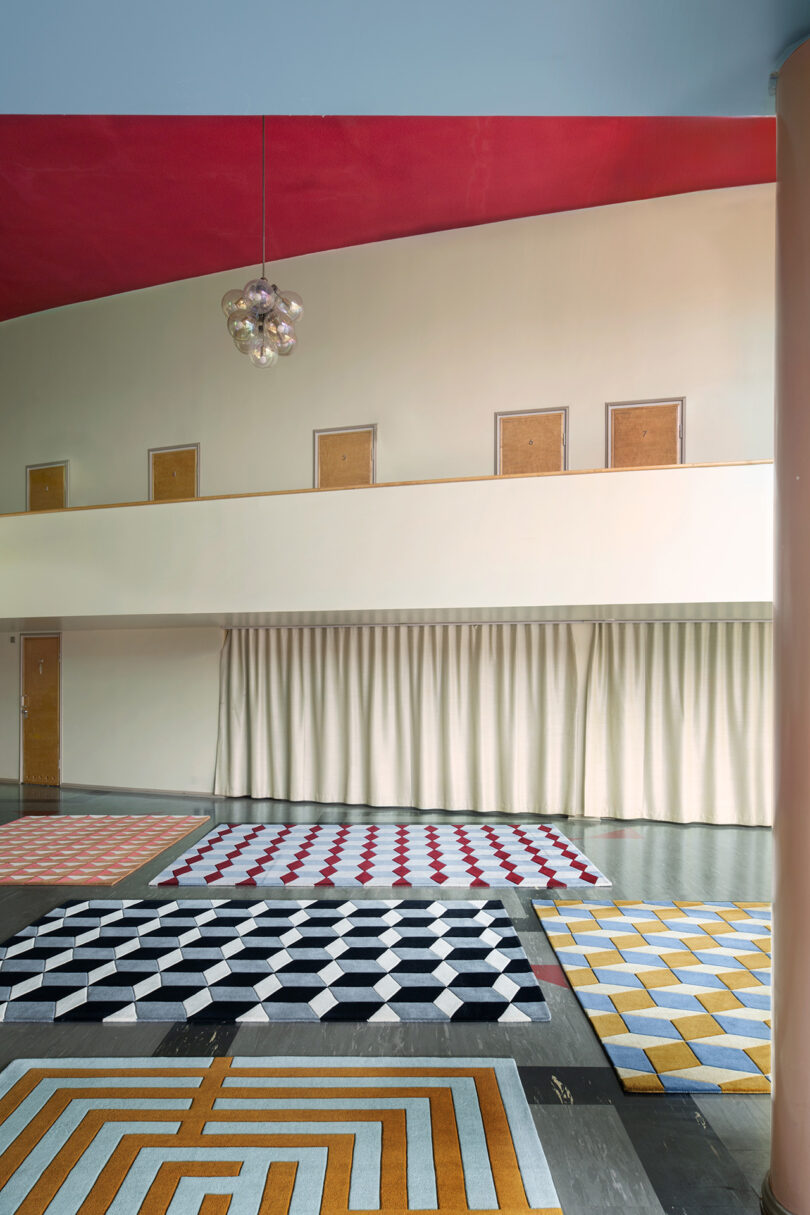 The interior features colorful geometric-patterned rugs gracing a dark floor, set against beige walls peppered with small rectangular windows. A red ceiling hovers above, showcasing a suspended art fixture that reveals layers of style and design, promising 10 years unfolded in modern elegance.