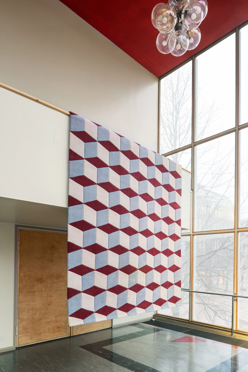 A large geometric-patterned textile from the "10 Years Unfolded" collection features striking red, blue, and white cubes. It elegantly hangs from a railing in a modern interior with tall windows and a bold red ceiling by LAYERED.