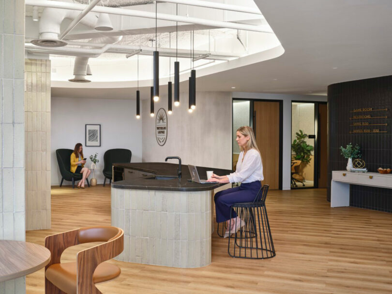 A woman works on a laptop at a modern office reception area with wooden floors and circular overhead lighting. Another person sits in the background.