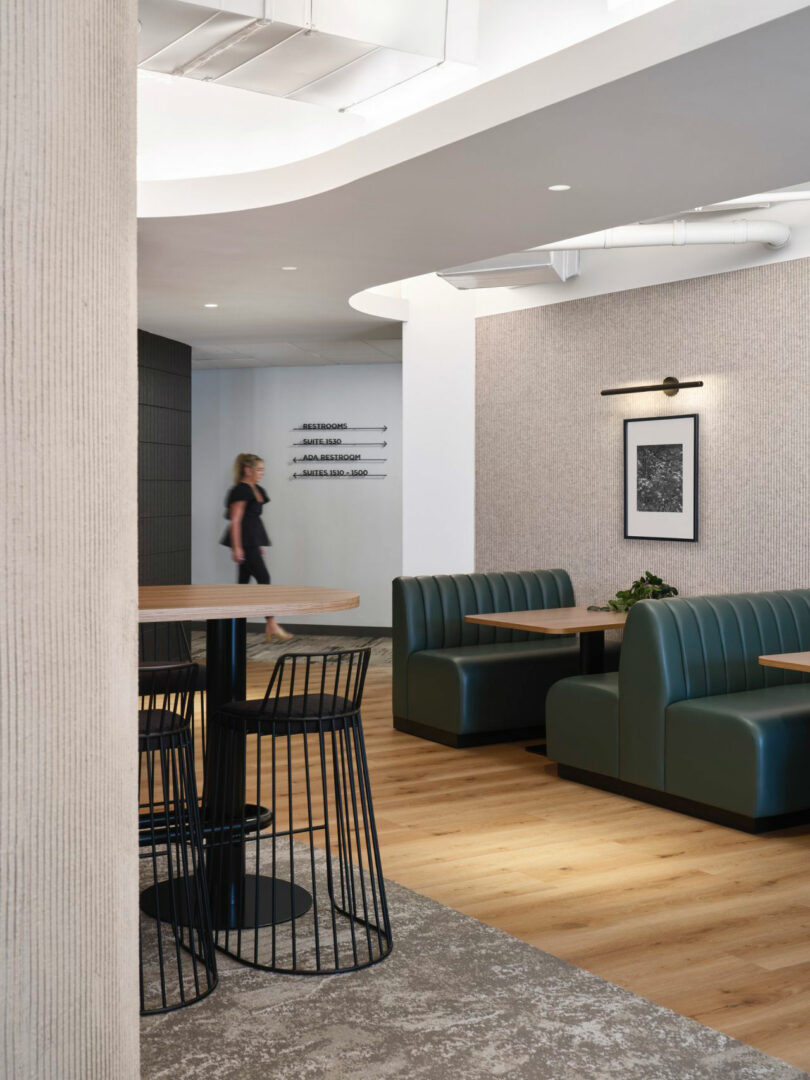 Modern cafe with green booth seating, a high table with stools, and artwork on the wall. A person walks in the background.