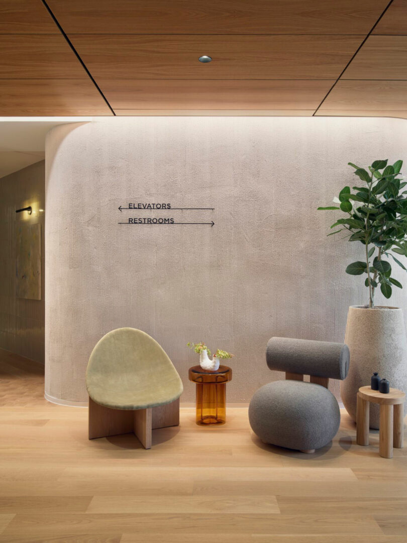 Modern lobby area with a curved wall, signage for elevators and restrooms, and minimal seating. A potted plant stands next to a small table with decor.