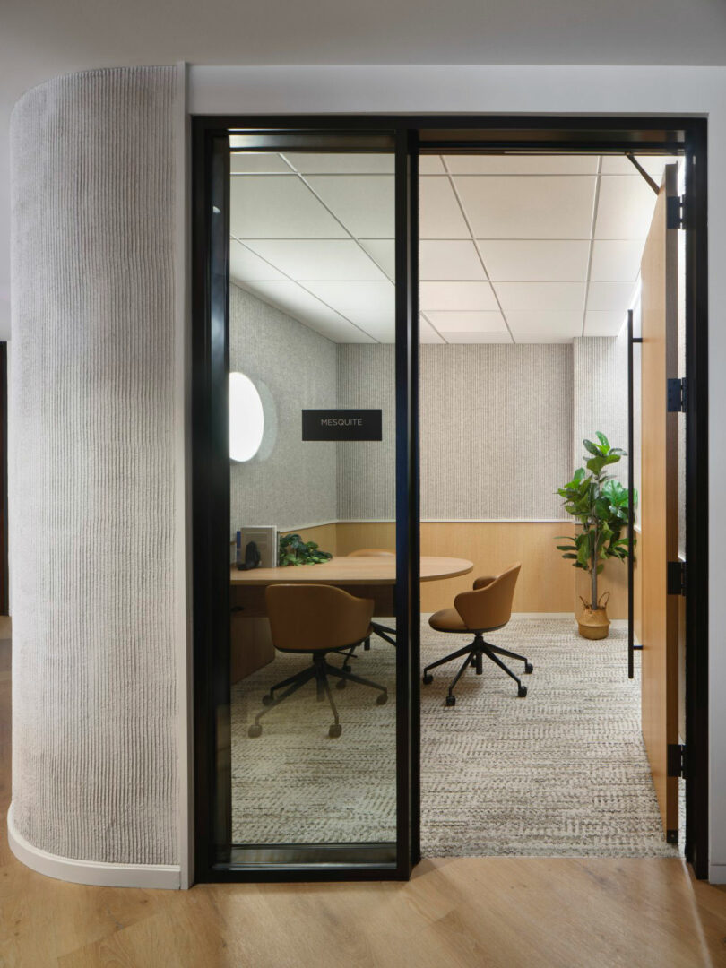 Small conference room with glass door, round table, and three brown chairs. Room has a textured wall, carpet, and a potted plant. Sign on door reads "Mesquite.