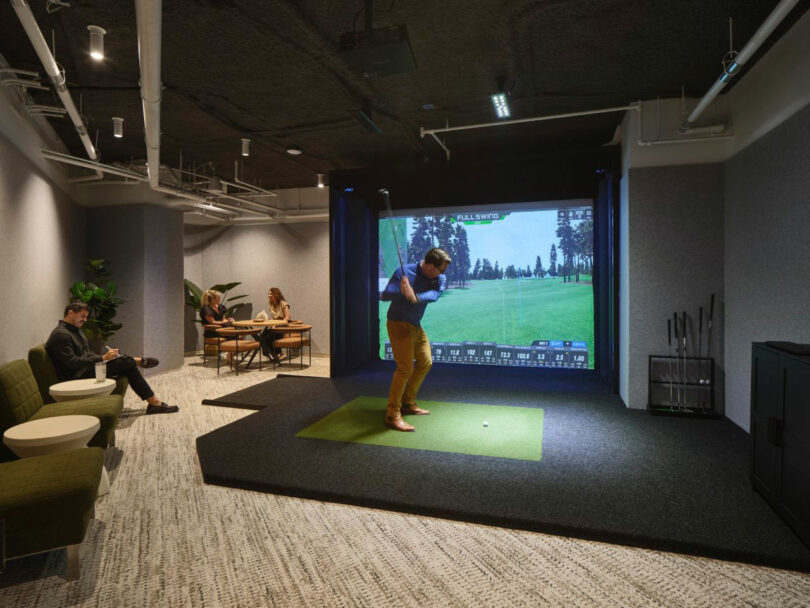 A person plays golf on a simulator in a modern indoor setup, while two others sit at a table in the background.