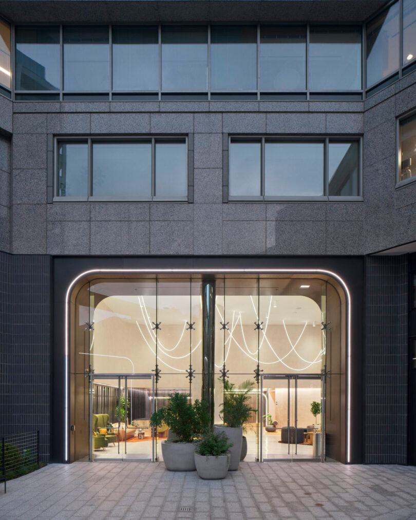 Modern building entrance with large glass doors, decorative lighting inside, and potted plants on the patio.