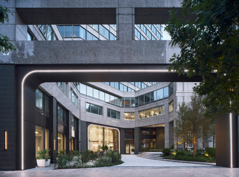 Modern office building with large glass windows, stone facade, and a curved entrance archway surrounded by greenery.