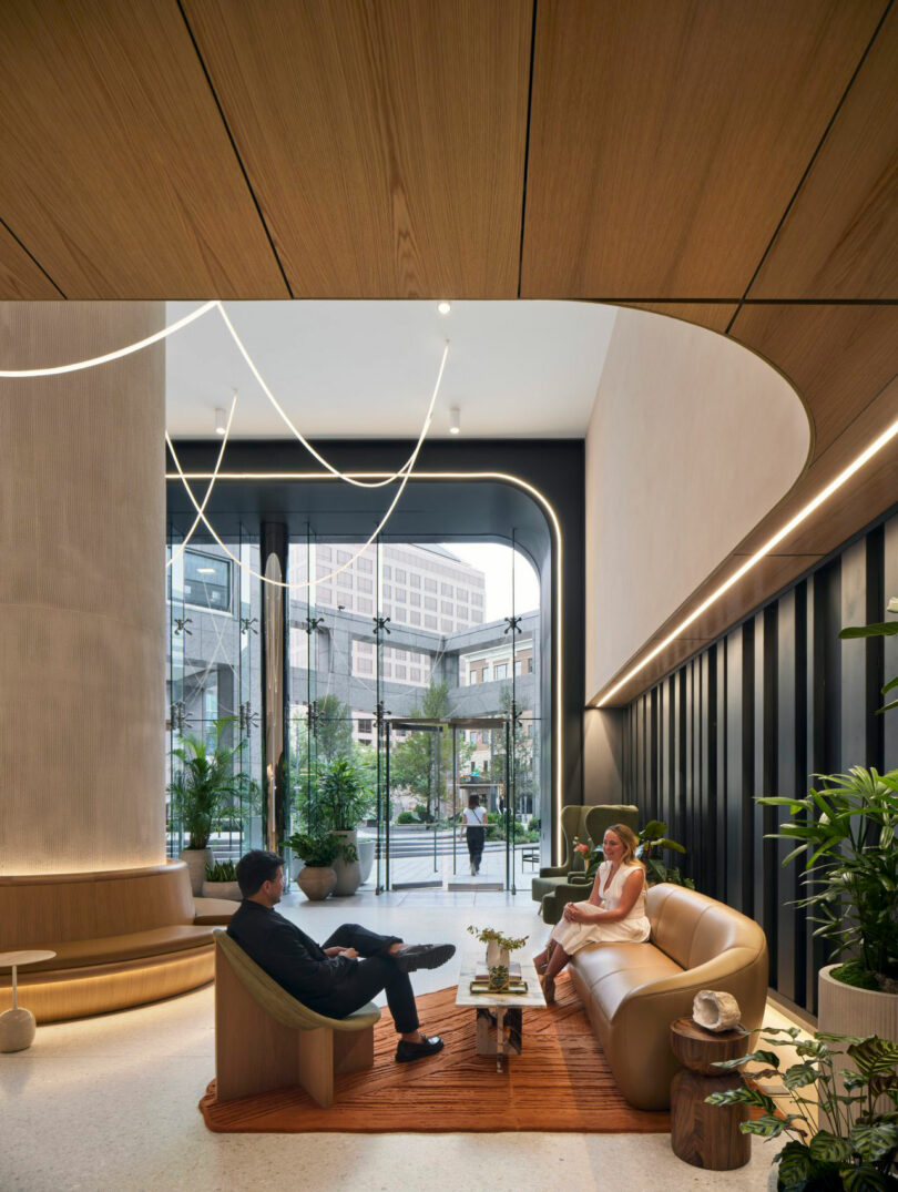 Two people sitting on a sofa in a modern lobby with large windows, plants, and curved wooden ceiling features. A city street is visible outside.