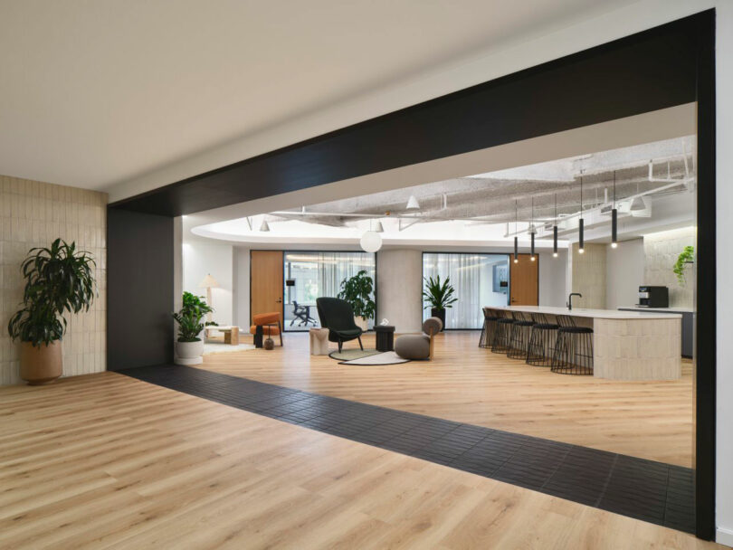 A modern open-plan office with wooden floors, a central lounge area with chairs, a long table with bar stools, and several potted plants.