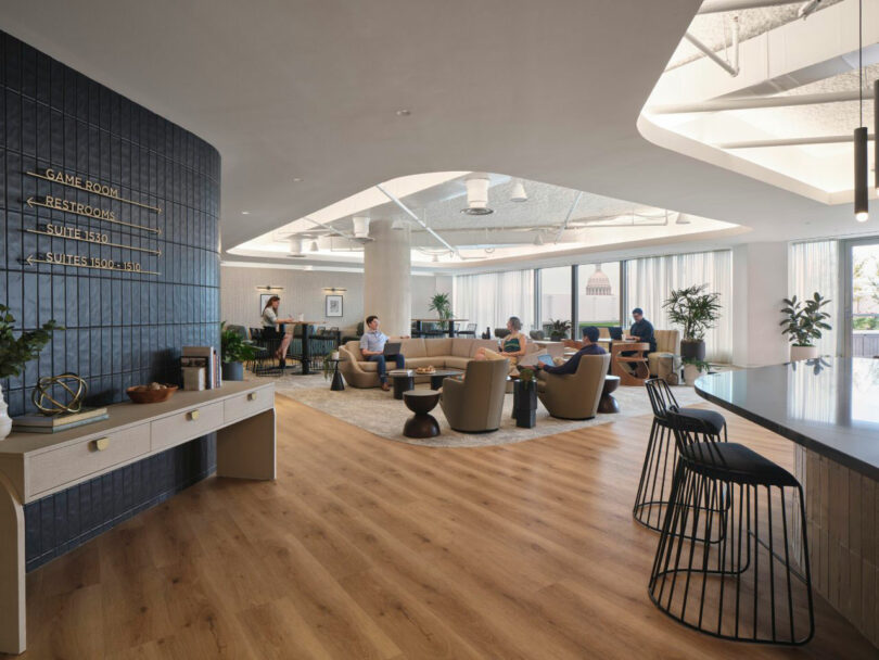 Modern office lounge area with people sitting and working on laptops. Features wooden floors, plants, and a sign pointing to the game room and restrooms.