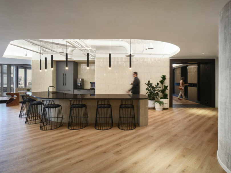 Modern office kitchen with a large island, bar stools, pendant lights, and a hardwood floor. A few people are walking around.