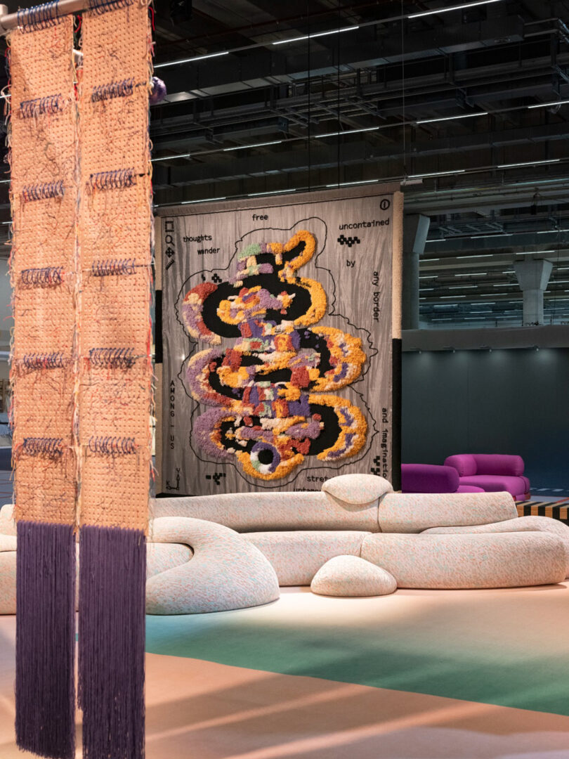 Modern art installation featuring a colorful abstract tapestry on a wall, textured columns, and curvy, multi-toned furniture on a light floor.