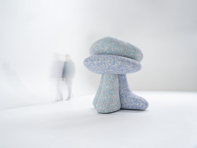 A large, abstract sculpture with soft, textured surfaces in muted colors is placed in a minimal white room. Blurred figures walk in the background.