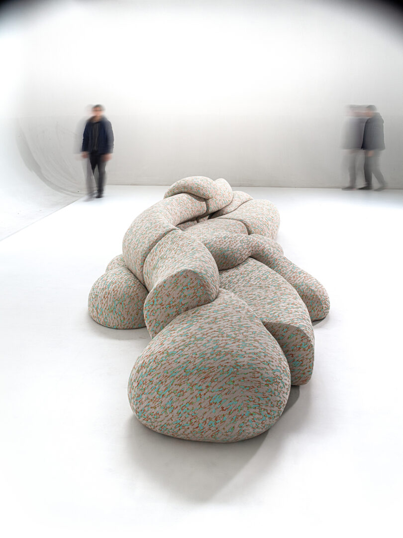 Sculptural seating arrangement resembling braided fabric with multicolored speckles, situated in a minimalist white room. Two blurred figures are walking by.