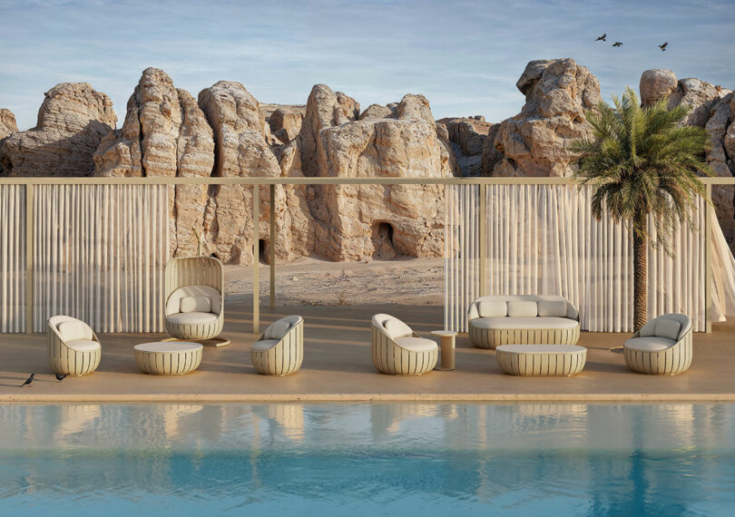 A modern outdoor seating area with armchairs and low tables is set by a pool, with rocky cliffs in the background and a few birds in the sky.