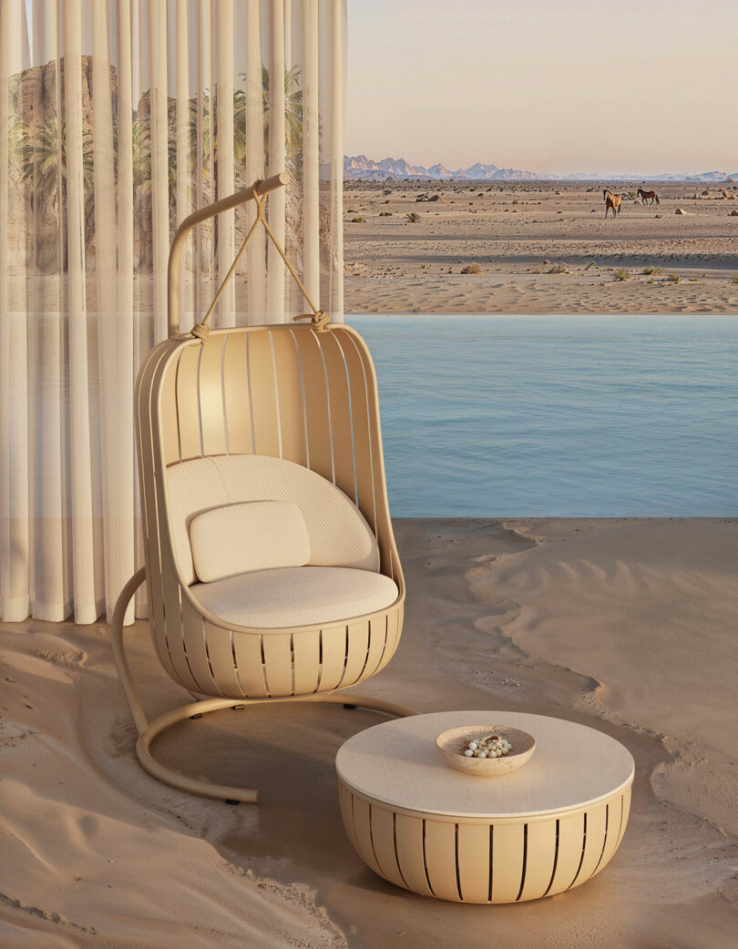 A beige hanging chair and round table on sandy ground by a water body, with sheer curtains and a desert landscape in the background.