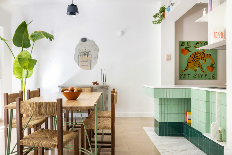 Bright dining room with a wooden table, woven chairs, and a large plant. Green-tiled fireplace on the right and modern art on white walls complete the decor.