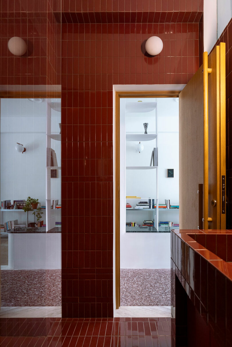 Interior view of a room with glossy red-tiled walls, a wooden door, and a partial view into an adjacent room with shelves and books.