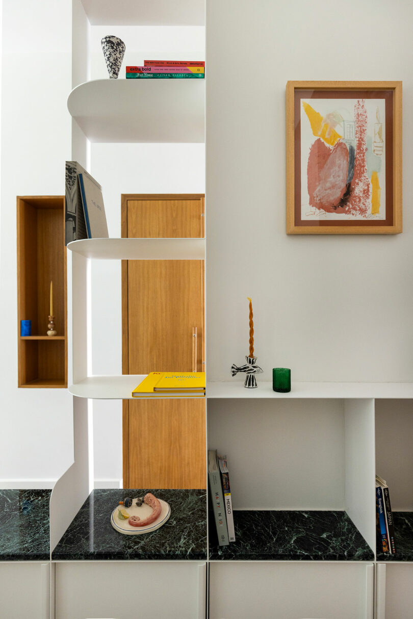 Modern interior with white shelves, geometric design, books, art, green marble base, and decorative items including a candle and framed painting.