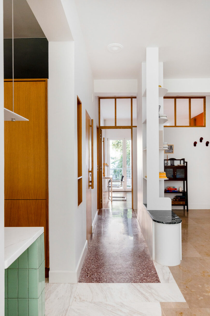 Modern hallway with a marble floor, white walls, and wooden accents. Shelving divides the space, leading to a well-lit room with a large window.