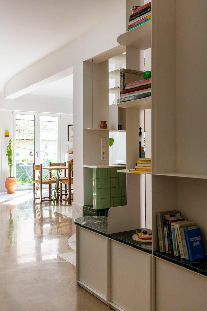 Modern interior with a bookshelf partition, green tile accents, a dining table with chairs by large windows, and sunlight streaming onto a polished floor.