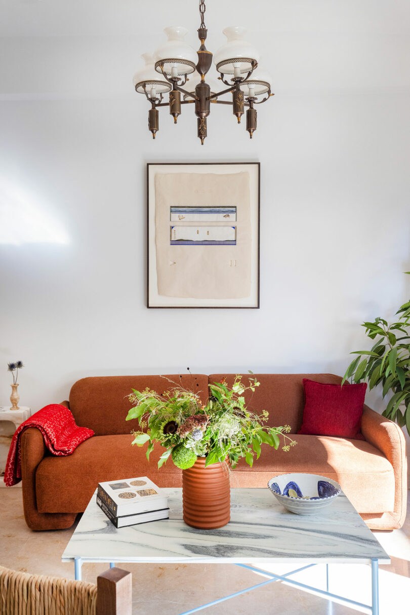 A cozy living room with an orange couch, red pillows, a marble coffee table, a plant, a chandelier, and artwork on the wall.