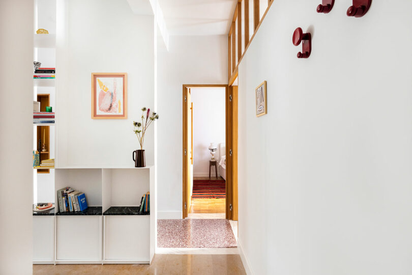 A bright hallway with white walls and shelving, featuring a few books and a vase. An open door leads to another room, and there are red hooks on the wall.