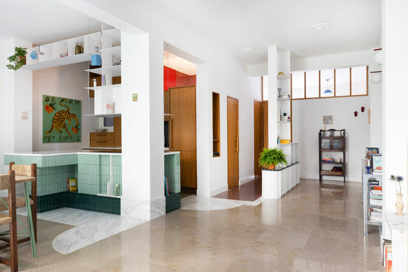 Bright, spacious room with white walls, green-tiled kitchen counter, and various decor. Bookshelf, plants, and furniture accent the modern interior. Marble and wood flooring visible.