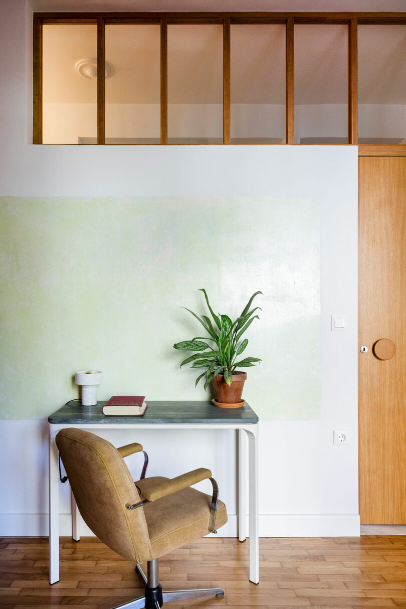 A small desk with a potted plant, a book, and a lamp is positioned against a light green wall. A brown swivel chair is in front of the desk. Wooden floor and partial wood-paneled upper wall visible.