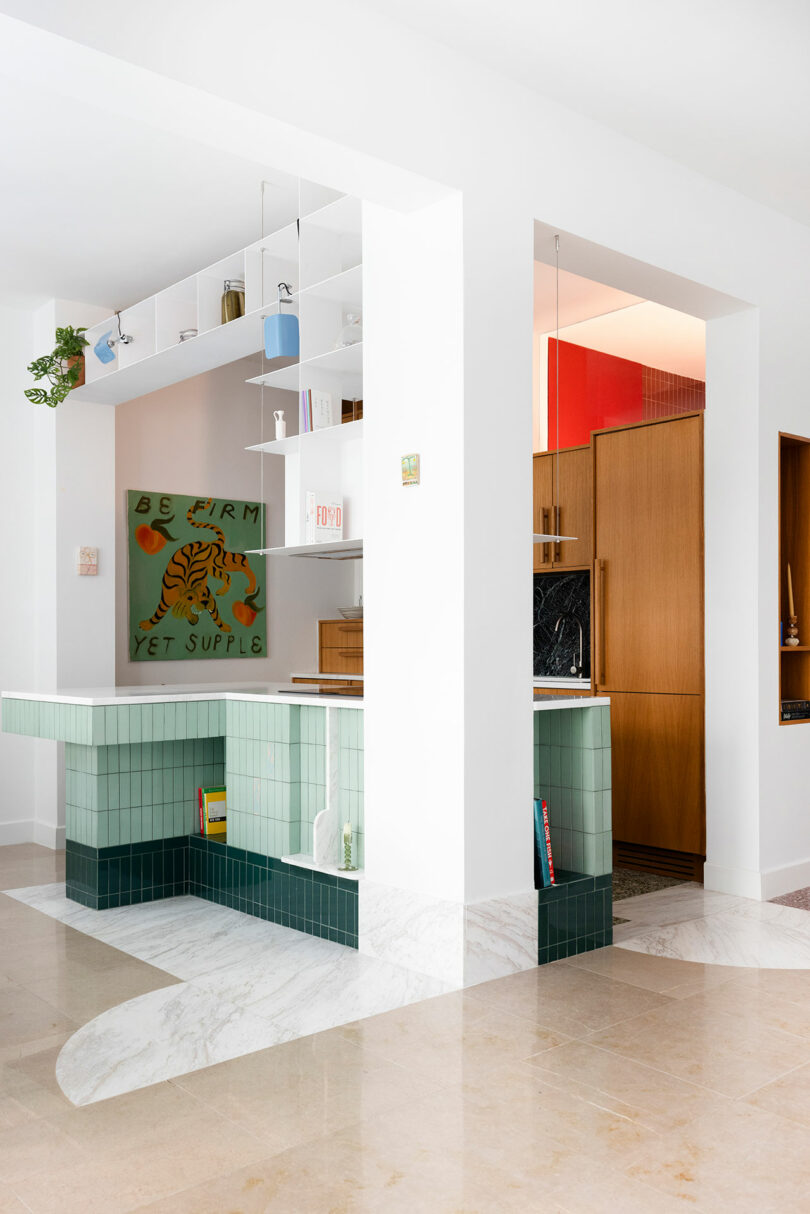 Modern kitchen with light green tiles and wooden cabinets. Open shelving holds books and decor. Wall art reads "Be firm yet supple" with a tiger illustration. Marble floor with geometric designs.