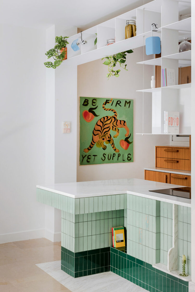 A modern kitchen with mint green and dark green tiles, white countertops, open shelving, and a wall art featuring a tiger with the phrase “Be Firm Yet Supple.”.