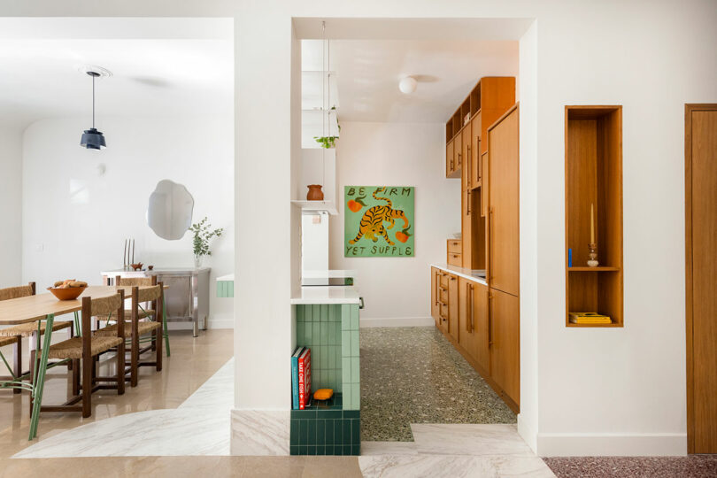 A modern kitchen and dining area with wooden furnishings, white walls, and green accents. A painting with the phrase "Be firm yet supple" featuring a tiger hangs on the wall.