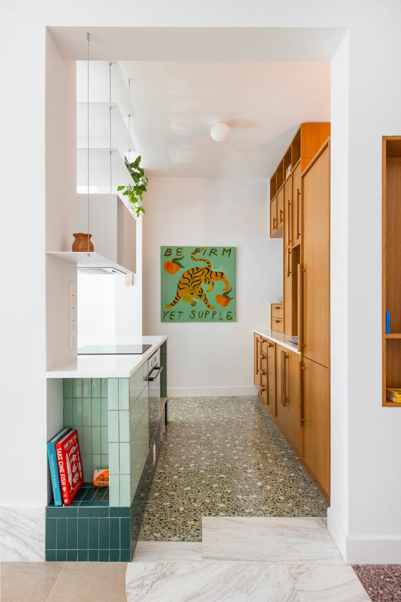 A modern kitchen with green tile, wooden cabinets, and terrazzo floor. A colorful artwork with a tiger and the phrase "Be firm yet supple" hangs on a white wall.