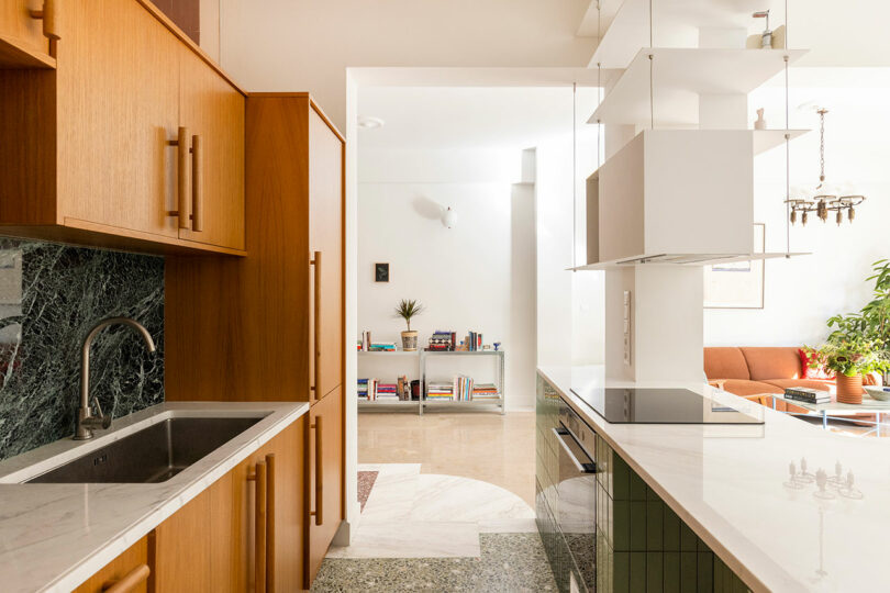 Modern kitchen with wooden cabinets, marble countertops, and green-tiled backsplash. An open doorway leads to a living area with a bookshelf and plants, visible in the background.