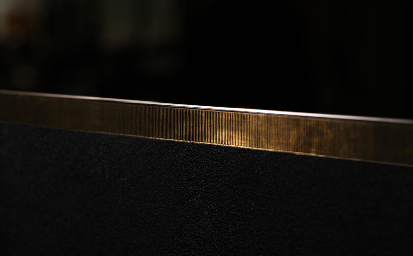 Close-up of a dark surface with a thin, golden metallic edge reflecting light. The background is out of focus, creating a contrast between the textured surface and the smooth metallic strip.