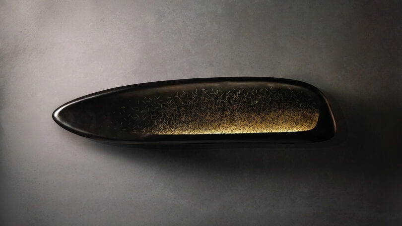 A sleek, elongated, black object with a reflective surface featuring a gradient of golden star patterns on the right side, set against a textured gray background.