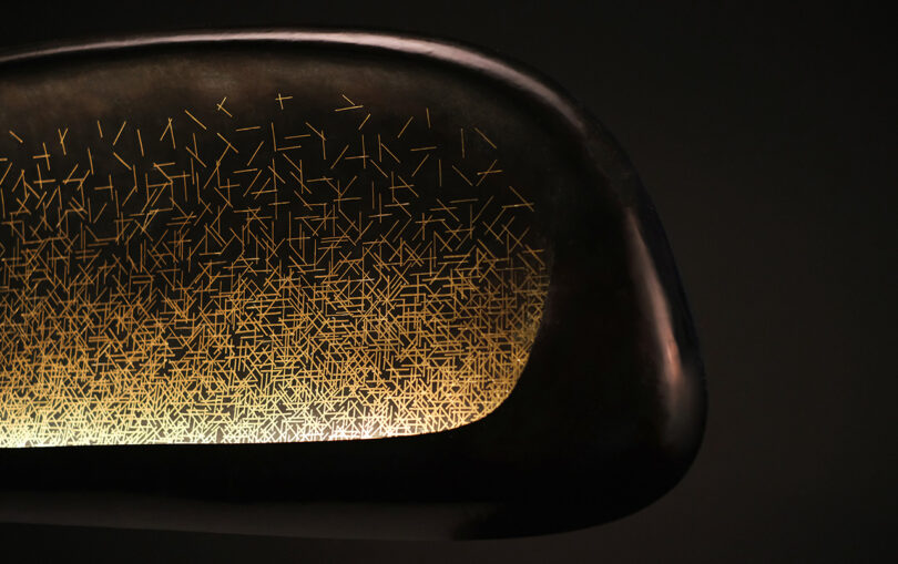 Close-up of a dark, curved object with intricate golden, linear patterns on its surface, set against a dark background.