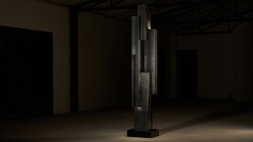 Tall, dark geometric sculpture stands illuminated in a dimly lit room with a visible beam structure on the ceiling.