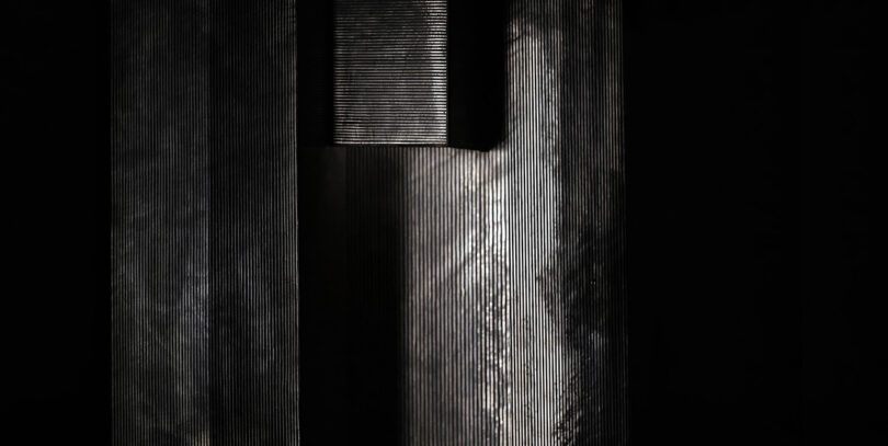 Vertical metal beams with textured surfaces cast contrasting shadows and light in a dark setting.