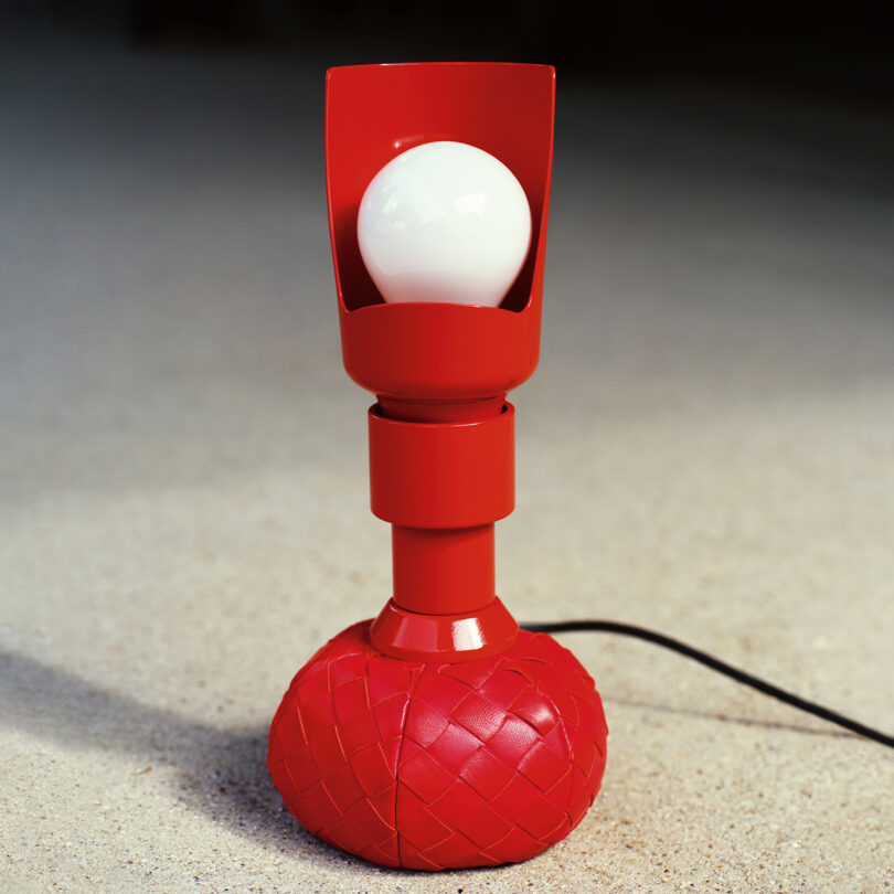 A red table lamp with a woven base and a visible light bulb on a neutral-colored surface.